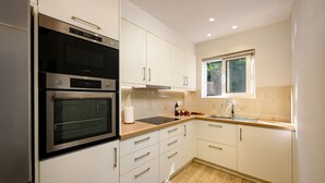 A modern kitchen with white cabinetry, a built-in oven, a microwave, and a window providing natural light. - Villa Idola Dassia Corfu (Corfu)