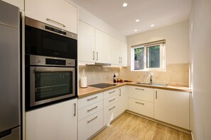 A modern kitchen with white cabinetry, a built-in oven, a microwave, and a window providing natural light. - Villa Idola Dassia Corfu (Corfu)