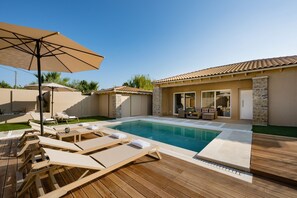 Piscina com espreguiçadeiras e um guarda-sol, rodeada por um deck de madeira e um muro de pedra.