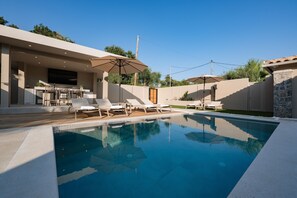 Une piscine entourée de chaises longues et de parasols, ainsi que d’un patio aménagé avec un téléviseur et du mobilier d’extérieur.