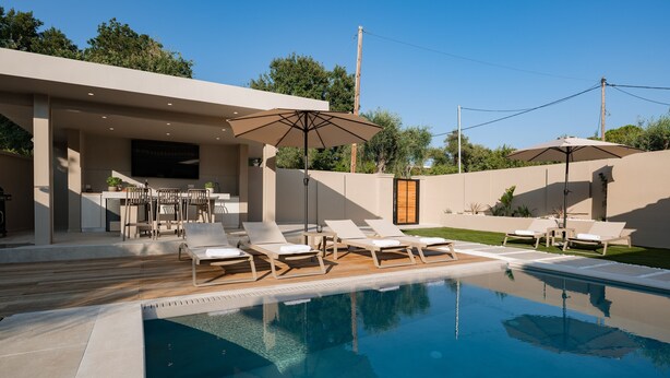 A modern outdoor pool area with lounge chairs and umbrellas, surrounded by a landscaped garden and a building with a flat roof. - Villa Idola Dassia Corfu (Corfu)
