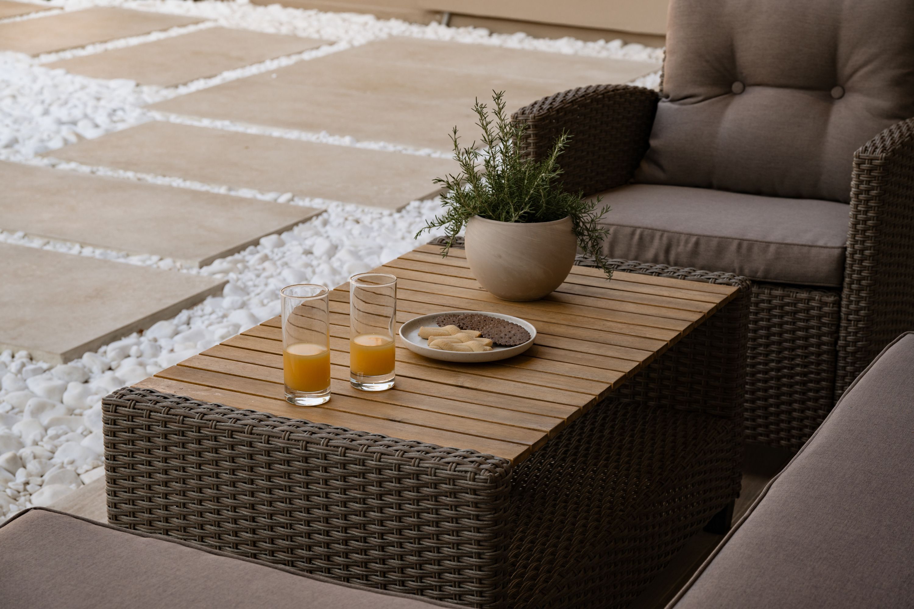 Ein Korbtisch mit zwei Gläsern Orangensaft und einem Teller Kekse, platziert auf einer Terrasse mit einem gewobenen Stuhl und einem Kissen.