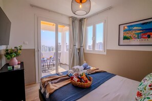 Une chambre d’hôtel avec un lit, une fenêtre donnant sur la ville, un balcon avec une table et des chaises, et un tableau accroché au mur.