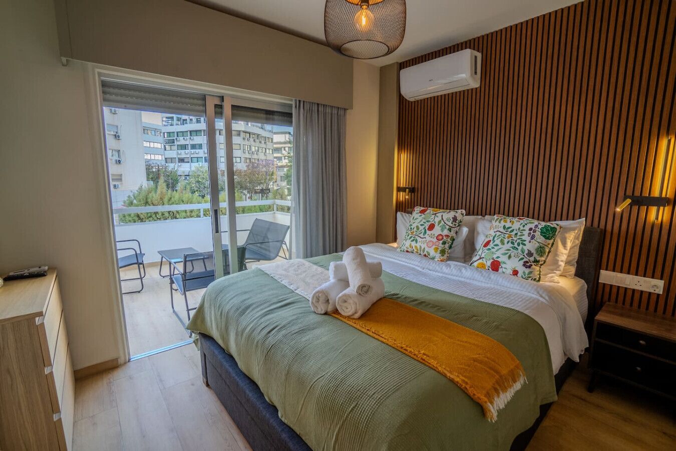 A hotel room with a bed, a desk, a chair, and a balcony with a view of buildings.