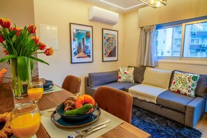 Ein modernes Wohnzimmer mit Essbereich, ausgestattet mit einer Sofagarnitur, einem Esstisch mit Obstschale und einer Vase mit Blumen.