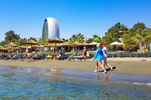 A beach with sun loungers and umbrellas, a tall building in the background, and palm trees. - 4.97 Super Host`s New Boutique & Prime Location (Limassol)