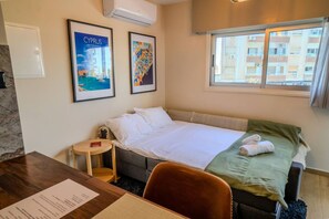 Une chambre d’hôtel avec un lit, une petite table, une chaise, une fenêtre donnant sur des immeubles et deux tableaux encadrés accrochés au mur.