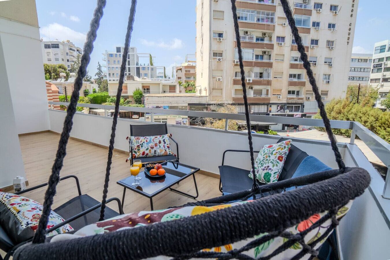 Een balkon met een hangstoel, een tafel met fruit en een glas sinaasappelsap, en uitzicht op woongebouwen.