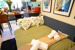 Ein gemütliches Zimmer mit einem Bett, Kissen, einem kleinen Tisch mit Früchten, einer Vase mit Blumen und einer gerahmten Karte an der Wand.