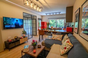 Ein modernes Wohnzimmer mit einer Couch, einem Couchtisch und einem Fernseher, der eine Sendung zeigt. Es gibt einen Essbereich mit Tisch und Stühlen sowie eine Küche mit einem roten Gerät.