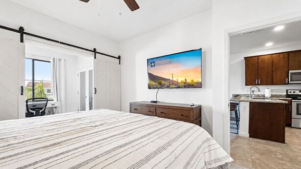 Quarto com cama, escrivaninha, cadeira, ventilador de teto, janela e vista para a cozinha.