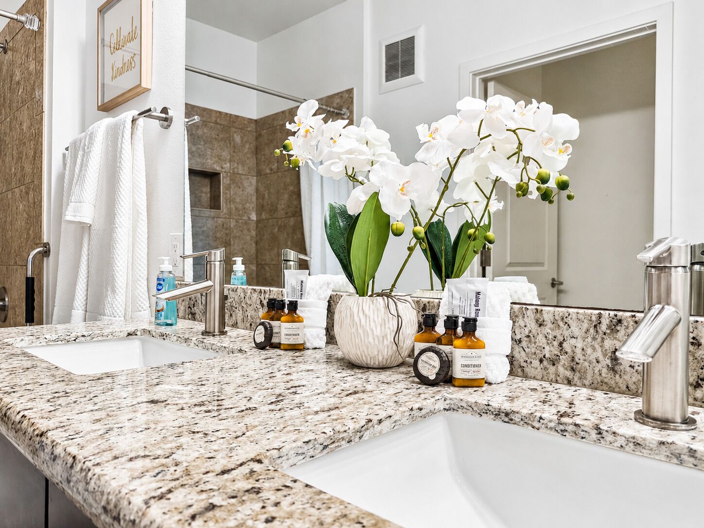 A bathroom with a granite countertop, two sinks, a mirror, a towel rack, and a vase with white flowers.