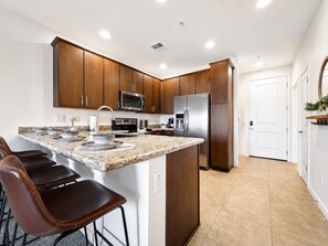 A modern kitchen with dark wood cabinets, stainless steel appliances, a granite countertop, and a bar stool. - Cays Luxury Living! Resort Pool, Gym, Walk to Restaurants (Chandler)