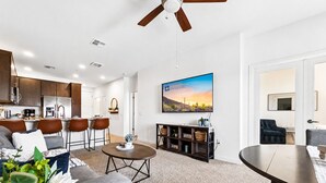 A modern living room with a sofa, coffee table, and TV, a kitchen with dark cabinets, and a ceiling fan.