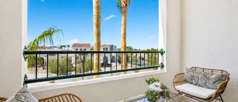 A balcony with wicker furniture, a glass table, and a view of palm trees and buildings.