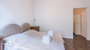 Un dormitorio con cama, mesitas de noche, lámpara y una puerta que da a otra habitación.