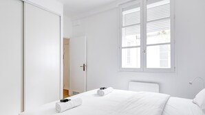 A minimalist bedroom with a large window, a bed with white linens, and a wardrobe. - BM – Élégance Parisienne & Vue sur Saint-Merri (Paris)