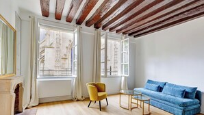 A room with a blue sofa, a yellow chair, and a small table. There are two windows with curtains, and a fireplace is visible on the left. - BM – Élégance Parisienne & Vue sur Saint-Merri (Paris)