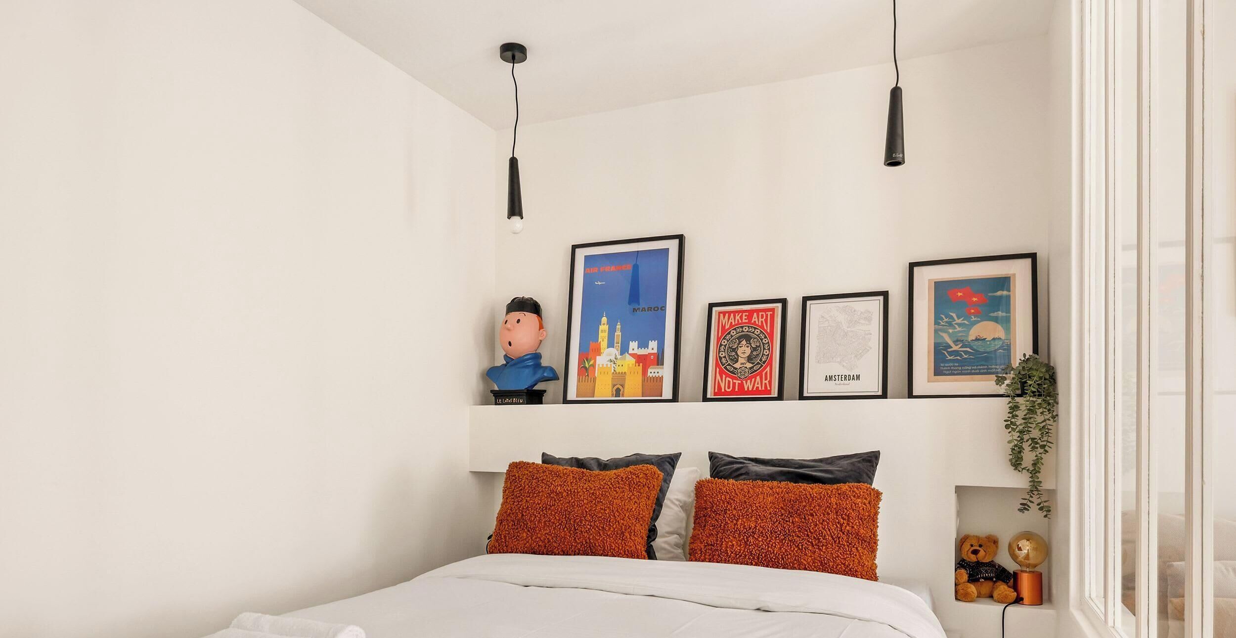 A bedroom with a bed, pillows, a wall-mounted shelf with framed artwork, and a hanging light fixture.