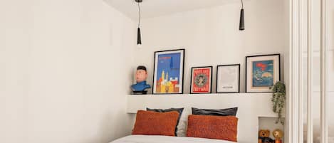 A bedroom with a bed, pillows, a wall-mounted shelf with framed artwork, and a hanging light fixture.