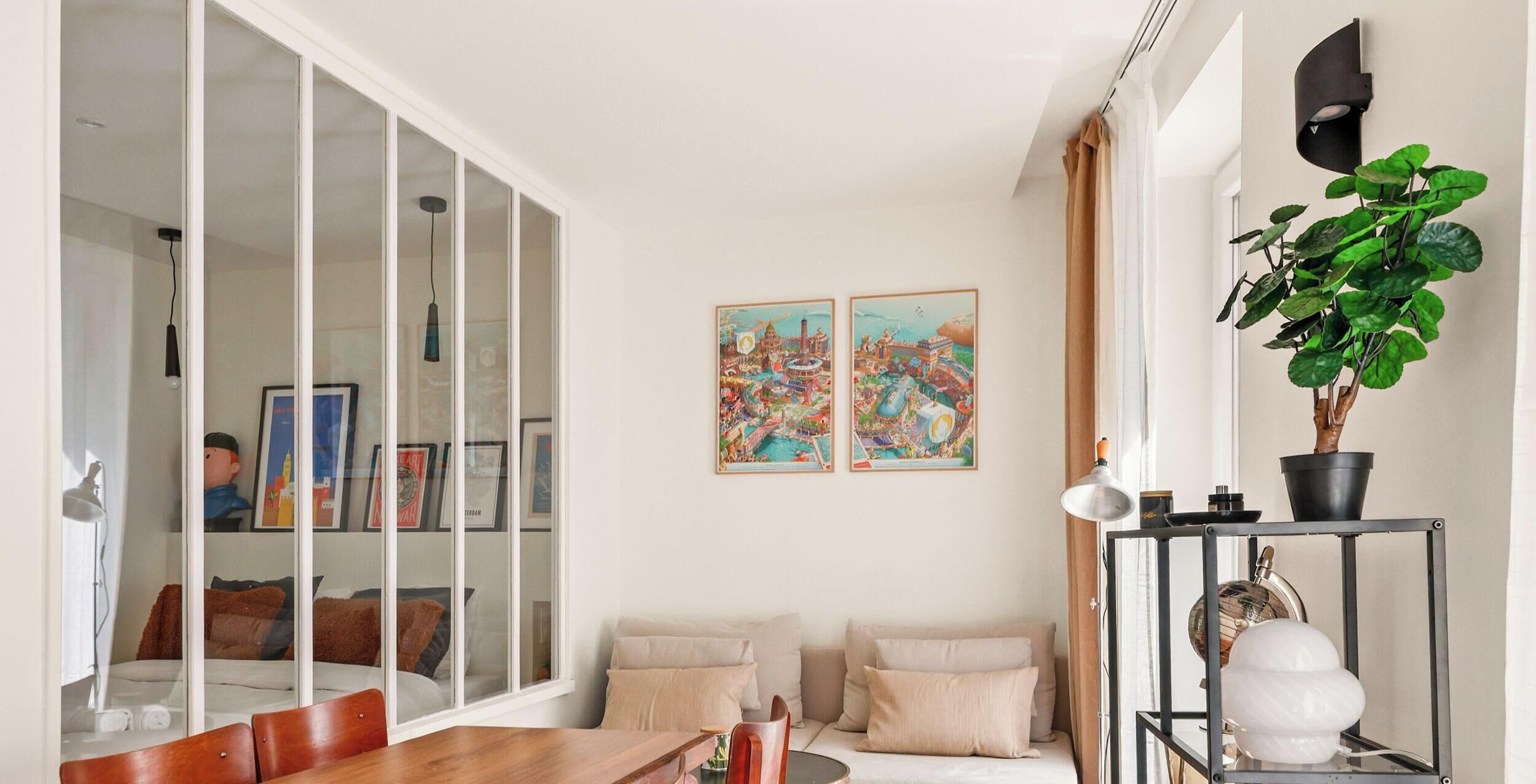 A modern living room with a glass partition, a dining area with a wooden table, and two framed artworks on the wall.