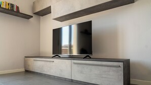 A modern living room with a flat-screen TV mounted on a grey cabinet, a built-in shelf with books, and a tiled floor. - Emilia Suite Holiday (Modena)