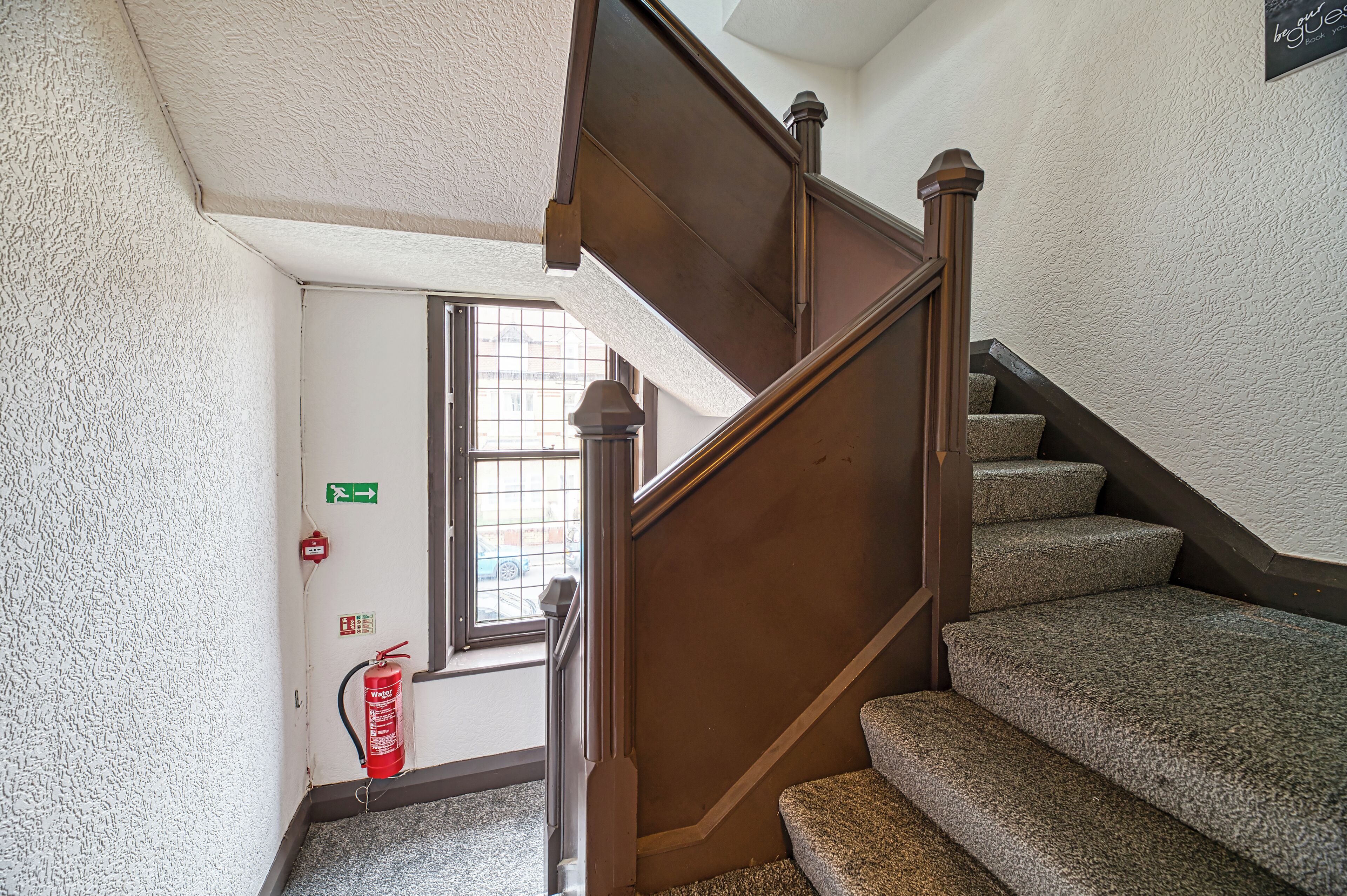 Escada com degraus carpetados e corrimão de madeira, levando a uma porta com janela e placa de saída de emergência.