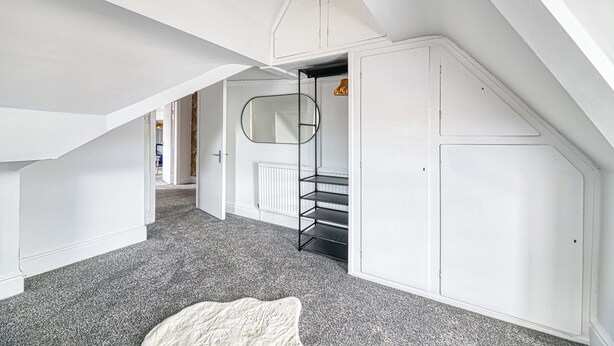 A room with a sloped ceiling, white walls, a grey carpet, a white door, and a metal shelf unit. - Glendale House Two Bedroom Apt By Guestz (Lytham St Annes)