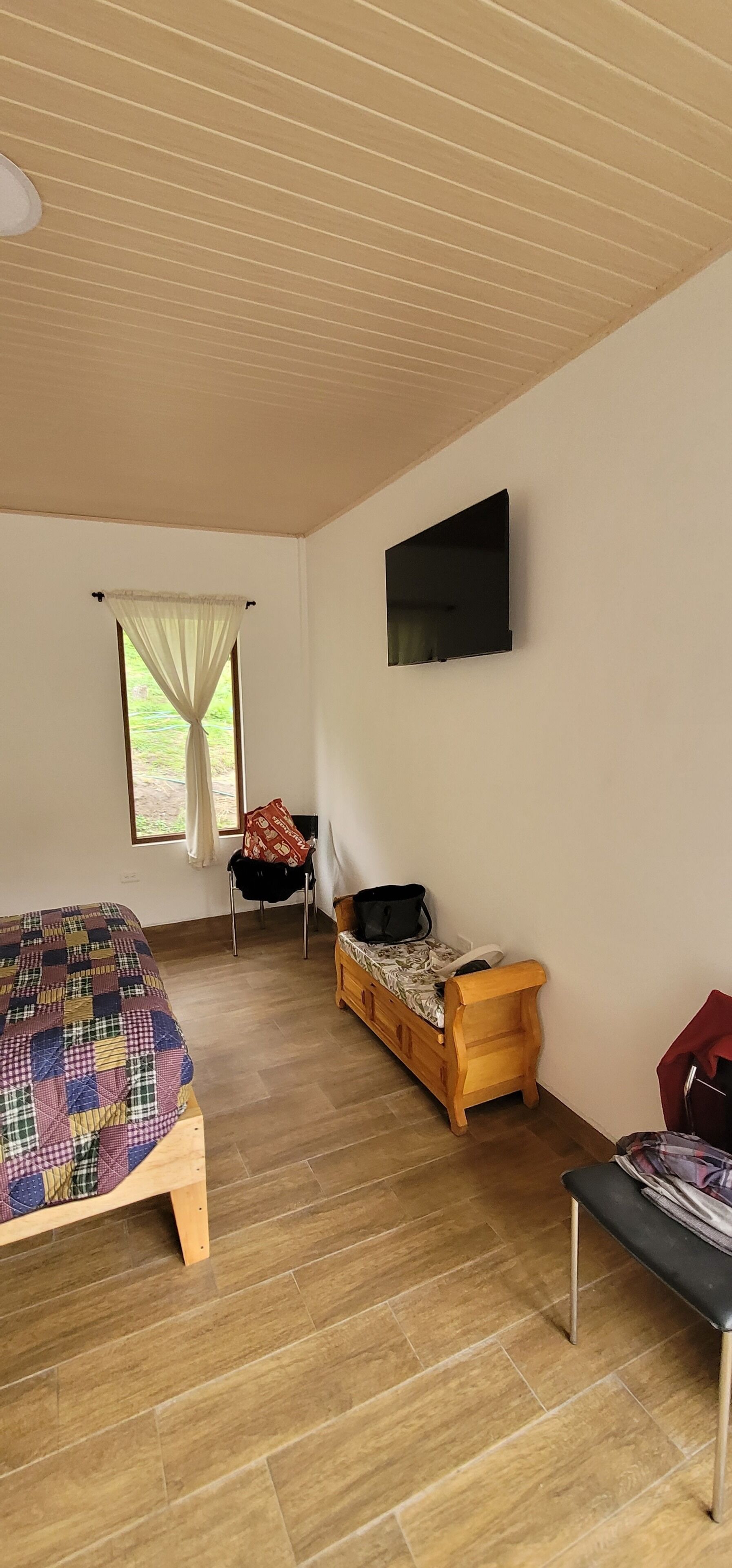 Ein Zimmer mit einem Bett, einer hölzernen Bank, einem kleinen Tisch, einem Fernseher und einem Fenster mit Vorhängen.