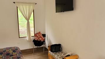 Ein Zimmer mit einem Bett, einer hölzernen Bank, einem kleinen Tisch, einem Fernseher und einem Fenster mit Vorhängen.