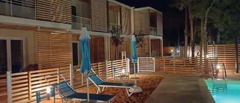 Zona de piscina con sillas de descanso y sombrillas, piscina y edificio con listones de madera y balcones.
