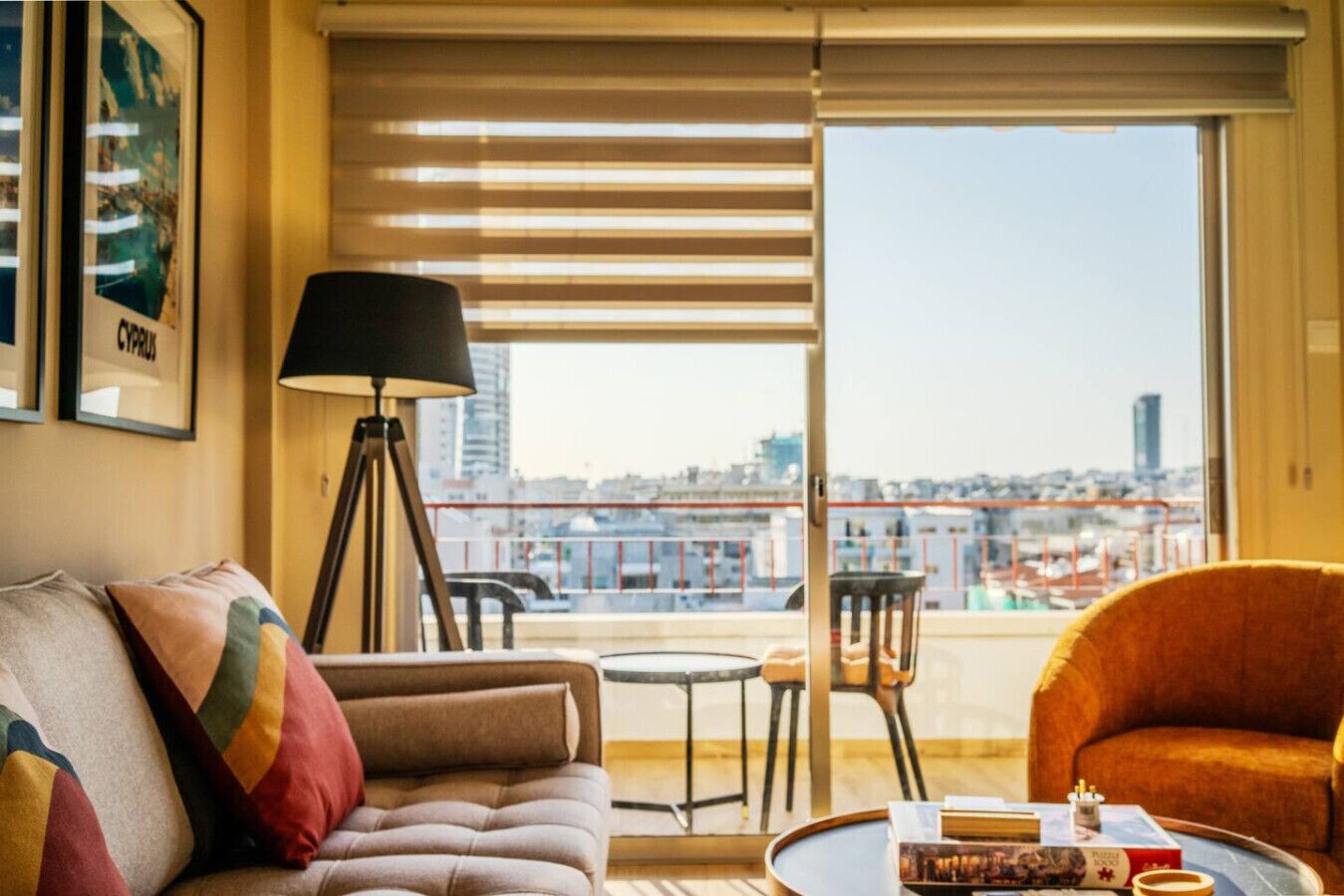A modern living room with a sofa, a lamp, a coffee table, and a view of a cityscape through large windows.