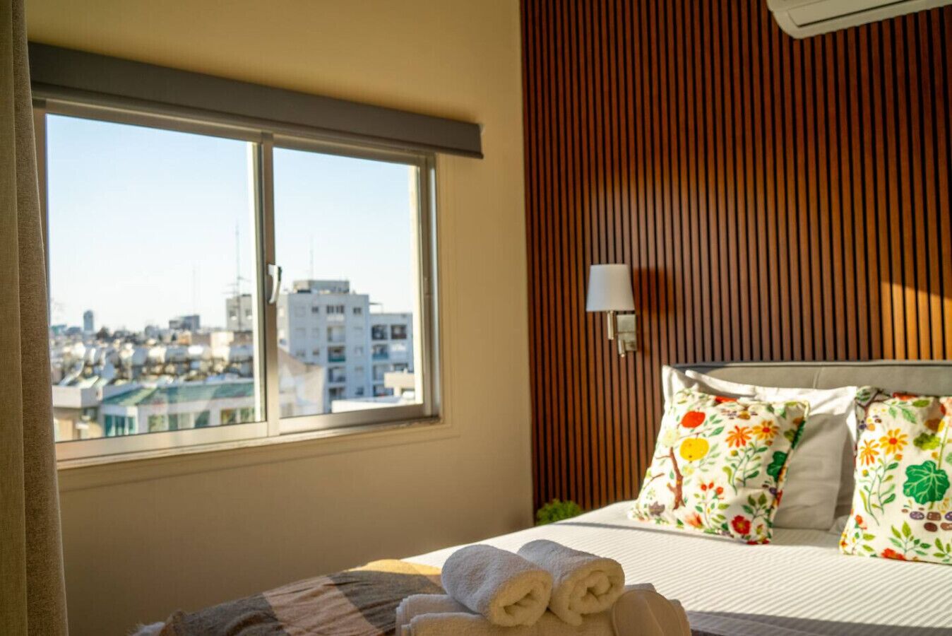 A hotel room with a large window, a bed with floral pillows, and a view of a cityscape.