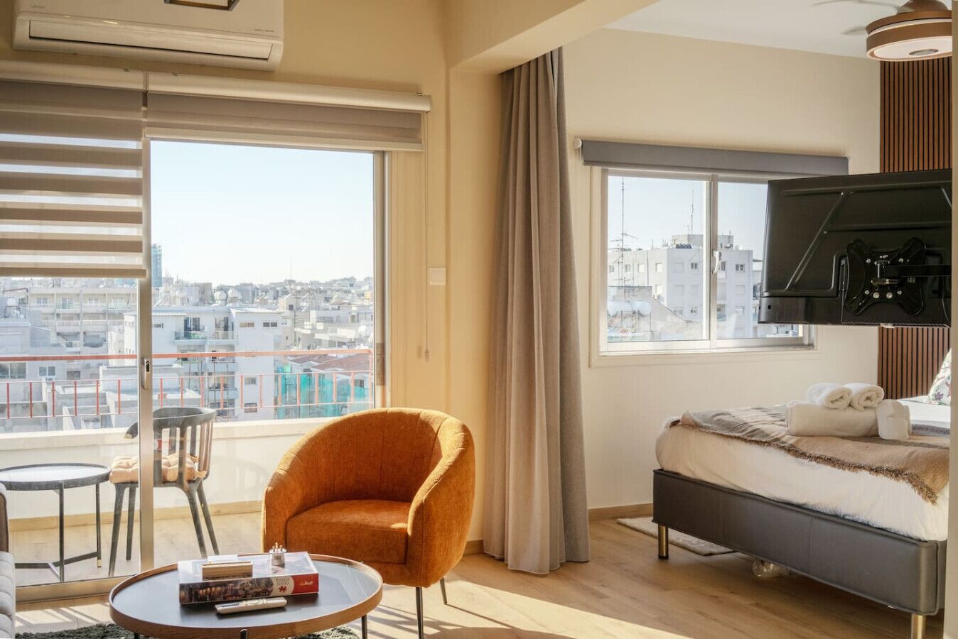 A hotel room with a bed, a chair, a table, a TV, and large windows with a city view.
