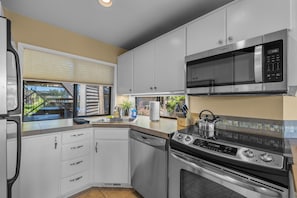 A compact kitchen with a microwave, stove, dishwasher, and a window with a view of a house and trees. - Mt Bachelor Village 332 | Hot tub | River Views (Bend)