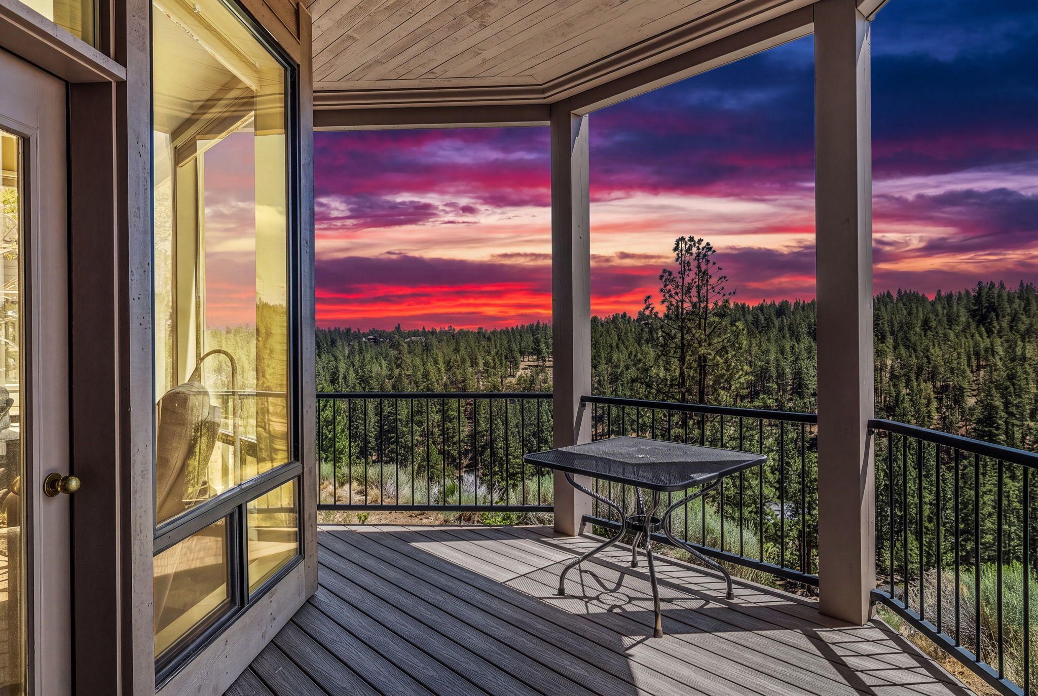 A wooden deck with a table and chairs, overlooking a forested area with a sunset view.
