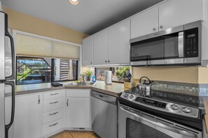 A compact kitchen with a microwave, stove, dishwasher, and a window with a view of a house and trees. - Mt Bachelor Village 332 | Unit A | Hot tub (Bend)