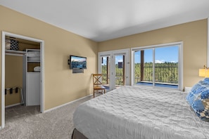 Quarto com cama grande, uma cadeira, televisão e varanda com vista para árvores.