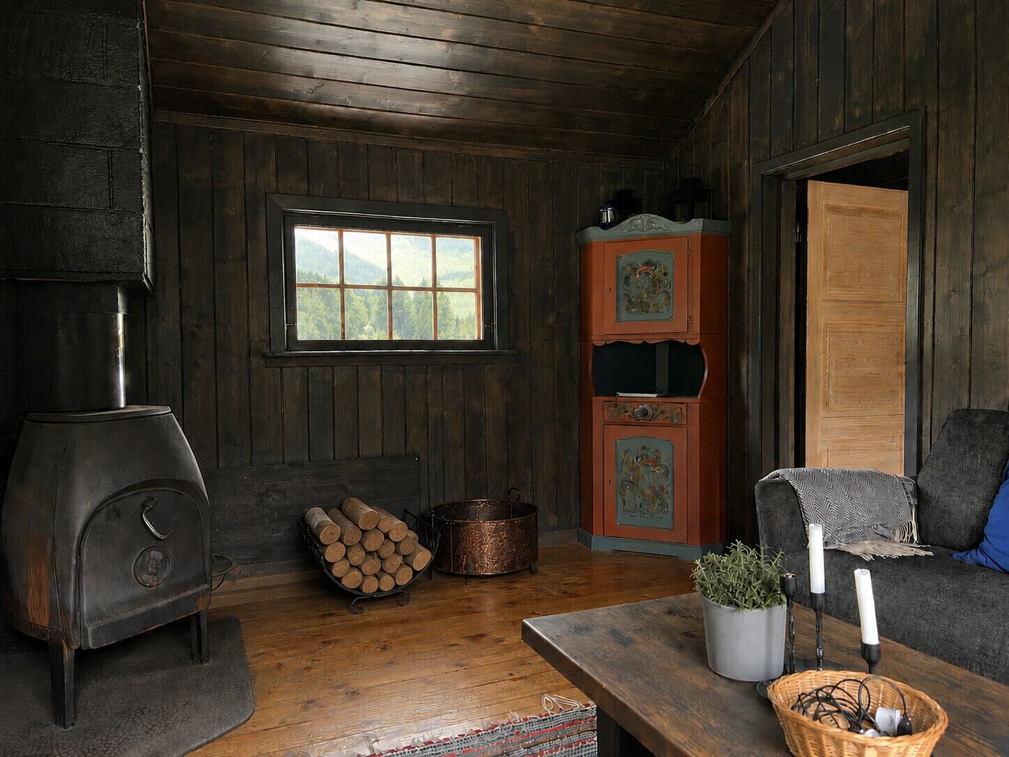Interior rústico de una cabaña con estufa de leña, ventana con vistas, un armario de madera y un rincón de estar acogedor.