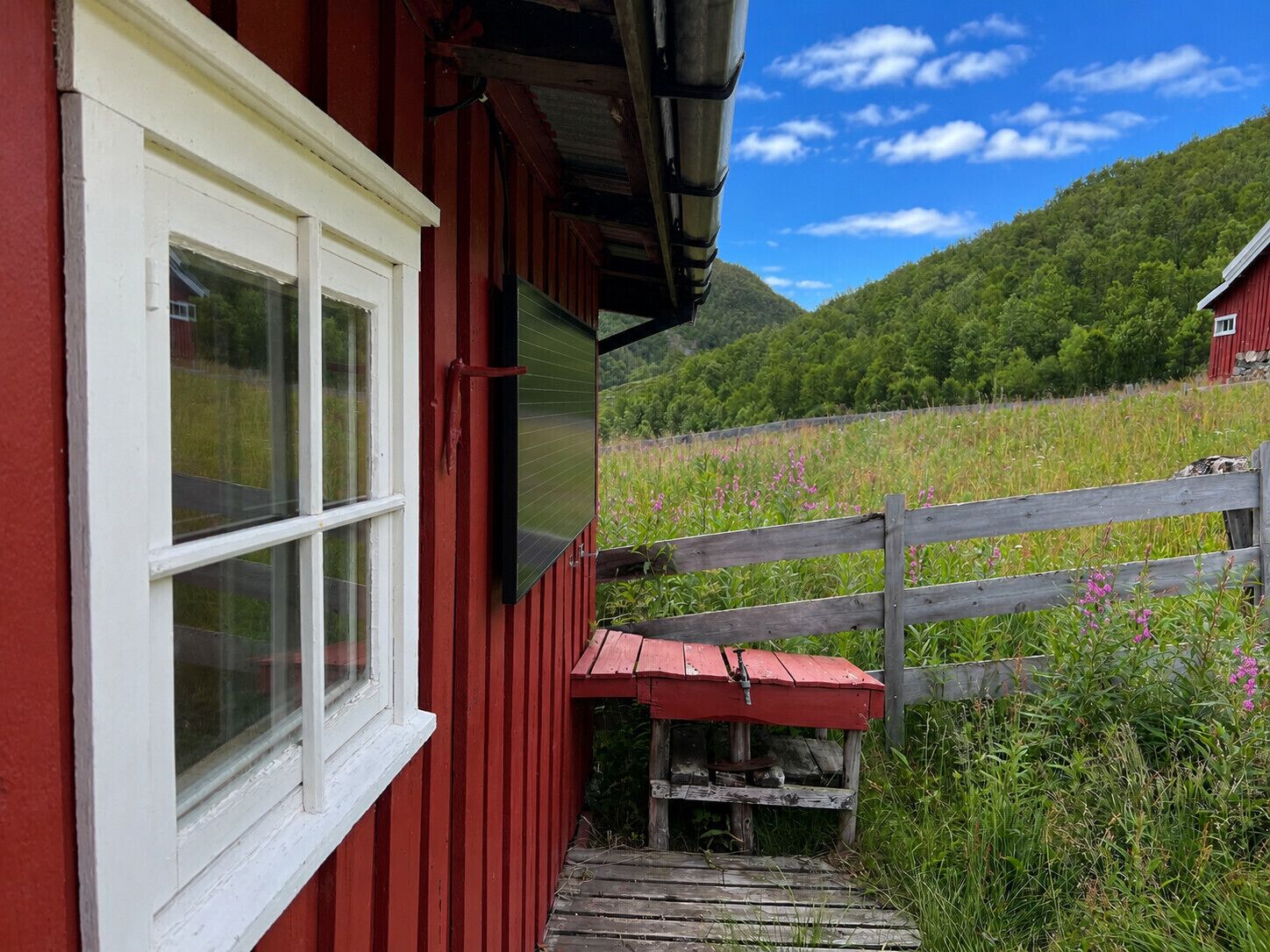 A red building with a white window, a wooden fence, and a view of a green field and mountains.
