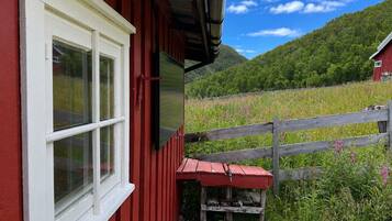 A red building with a white window, a wooden fence, and a view of a green field and mountains.