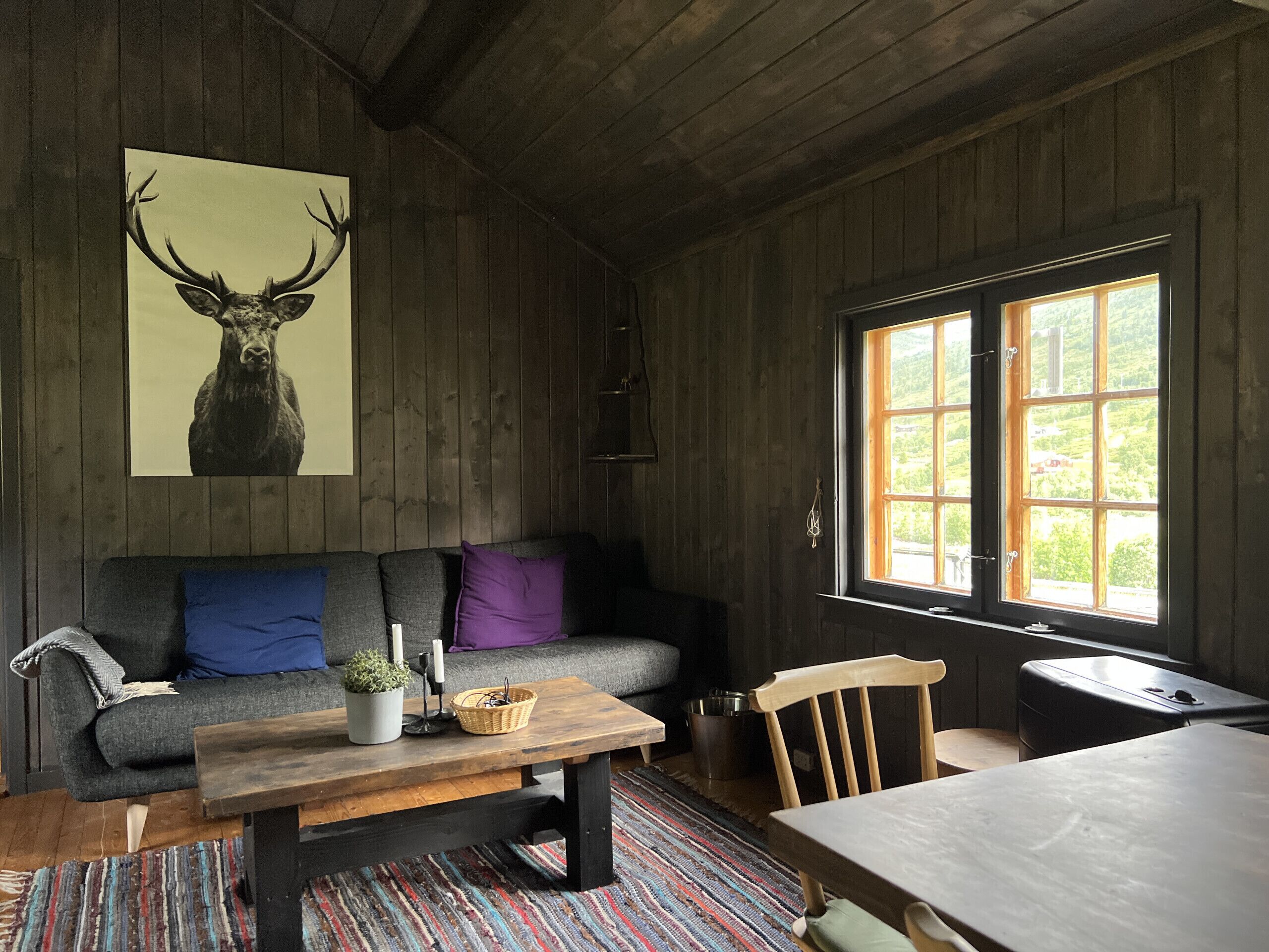 Una habitación acogedora con techo de madera, un sofá con cojines, una mesa con una cesta, una silla y una ventana con vistas.