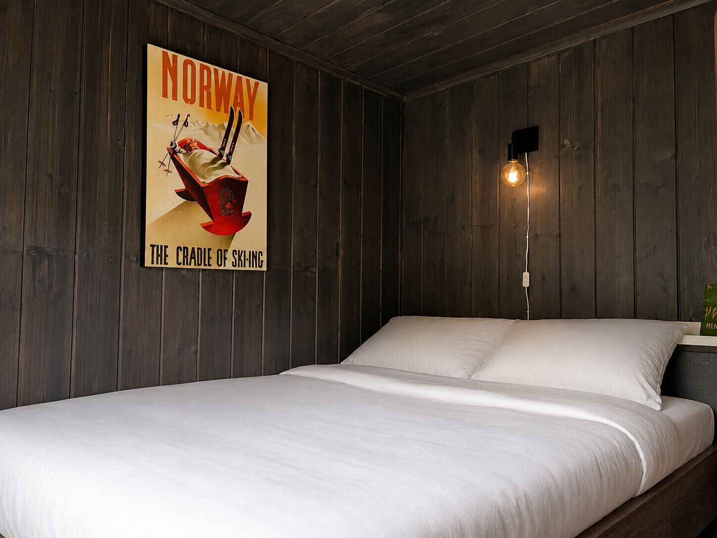 Ein Zimmer mit einem Bett, Holzwänden, einem Norwegen-Plakat und einer Wandleuchte.