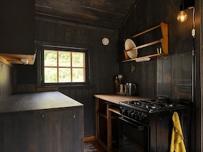 A kitchen with wooden walls, a window, a stove, and a hanging light fixture. - A Charming Off-Grid Cabin – Close to Nature in Hemsedal (Hemsedal)