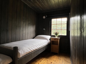 Ein kleines Zimmer mit einem Einzelbett, Holzboden und -wänden, einem Fenster mit Blick auf Grünflächen und einem Nachttisch mit Blumen in einer Vase.