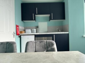 A dining area with a table and chairs, a kitchen in the background with cabinets and appliances, and a door to the left. - Aaron Wise - 09 Coldstream Studios (Cardiff)