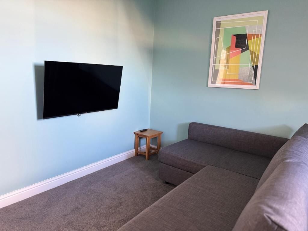 A living room with a flat-screen TV mounted on the wall, a small wooden side table, and a sectional sofa.