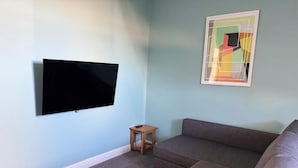A living room with a flat-screen TV mounted on the wall, a small wooden side table, and a sectional sofa. - Aaron Wise - 09 Coldstream Studios (Cardiff)