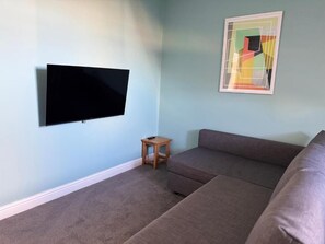 A living room with a flat-screen TV mounted on the wall, a small wooden side table, and a sectional sofa. - Aaron Wise - 09 Coldstream Studios (Cardiff)