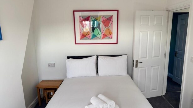 A hotel room with a bed, a small table, a framed artwork on the wall, and a door. - Aaron Wise - 09 Coldstream Studios (Cardiff)